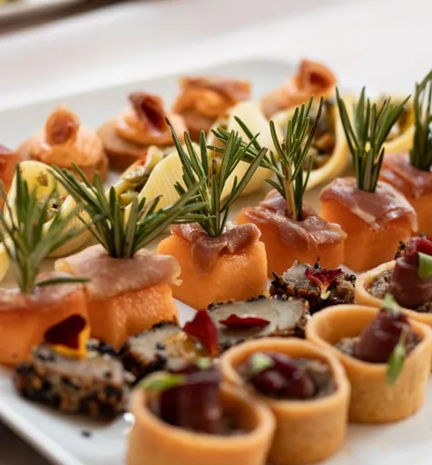 Bouchées de melon et jambon piquées de romarin, tartelettes et pièces cocktail sur plateau blanc