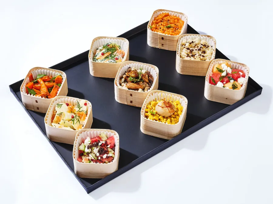 Barquettes individuelles en bois garnies de salades et de plats, sur plateau noir