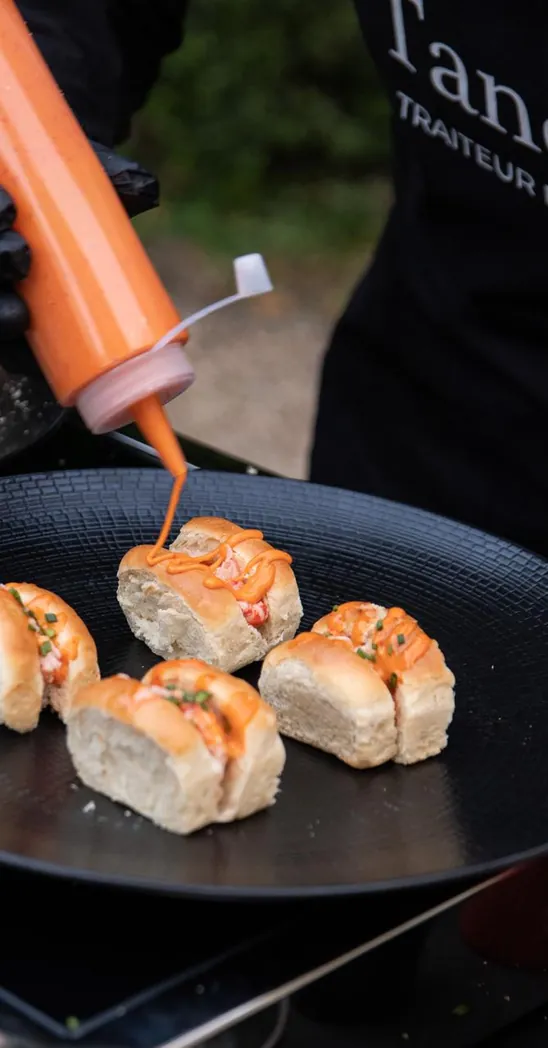 Cuisinier en tablier Tandrine nappant de sauce des petits pains garnis, sur une plancha